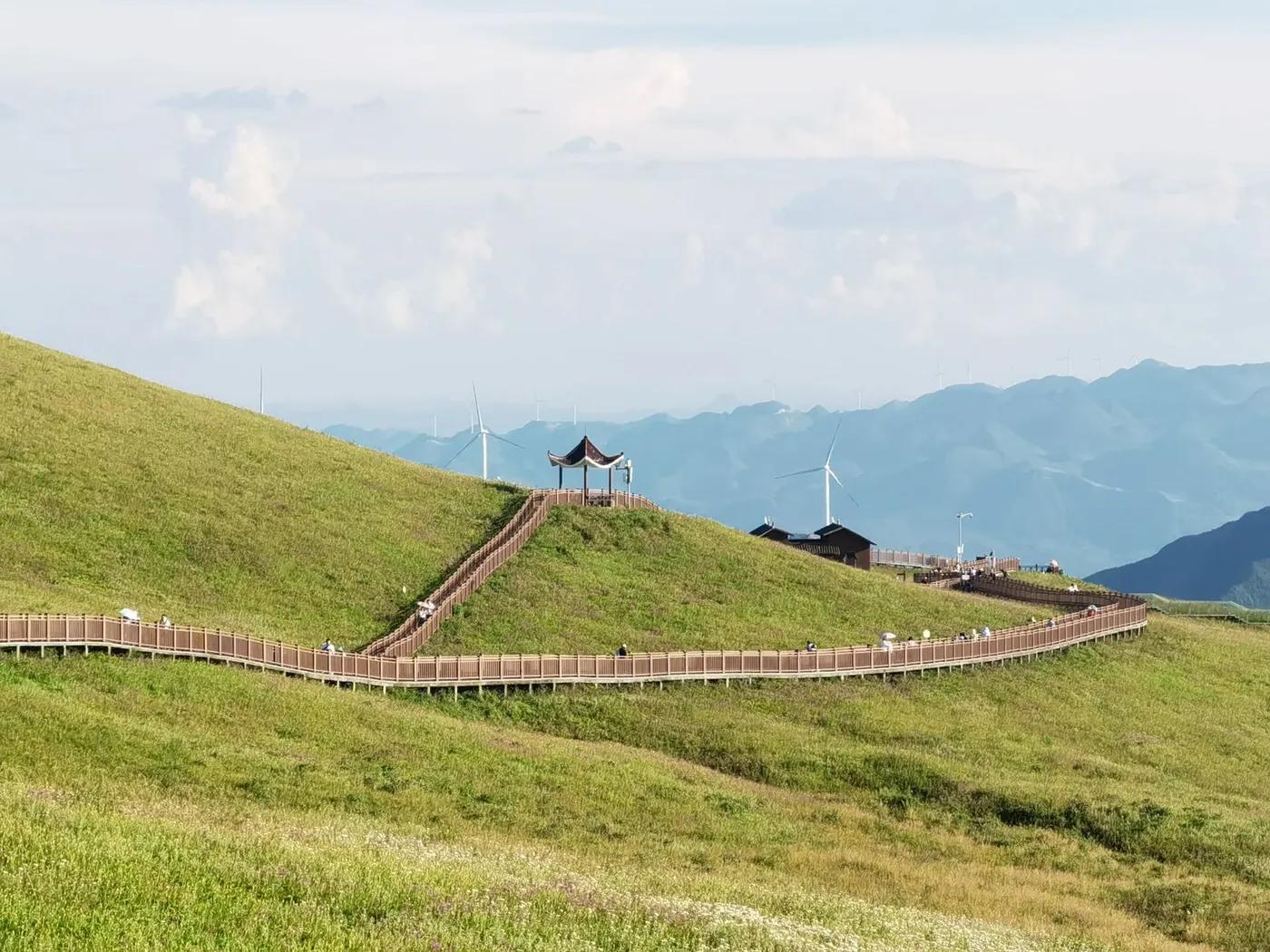 A picturesque pathway meanders through green hills with wind turbines in the distance, under a clear sky.