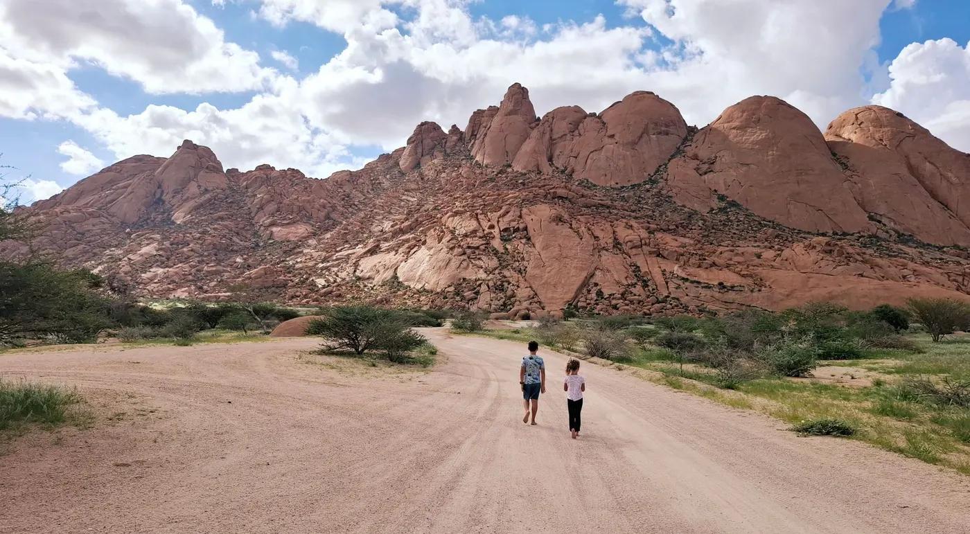 Two hikers walk along a trail under a vast Namibian rocky landscape.