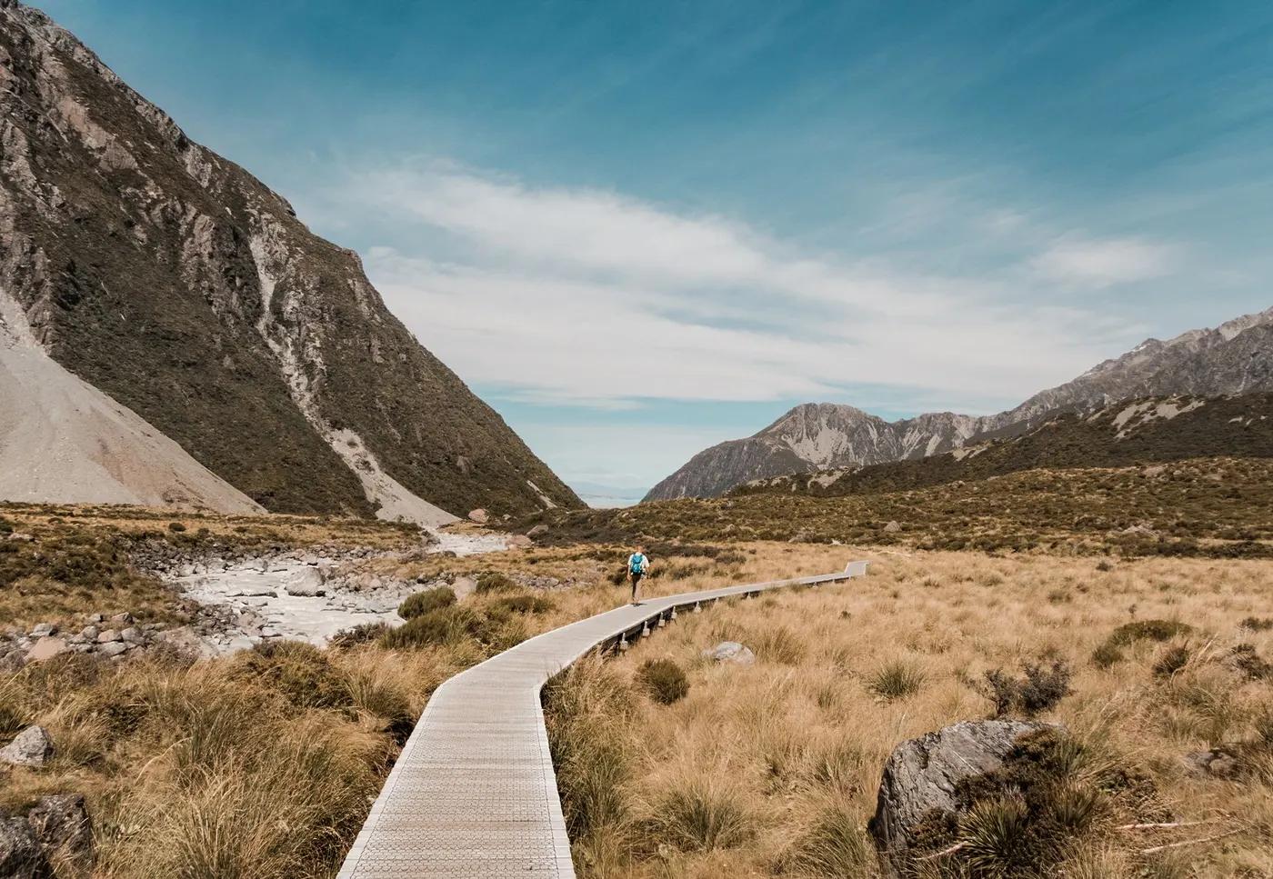 A hiker on a tranquil boardwalk path in New Zealand's mountainous landscape under a clear sky.