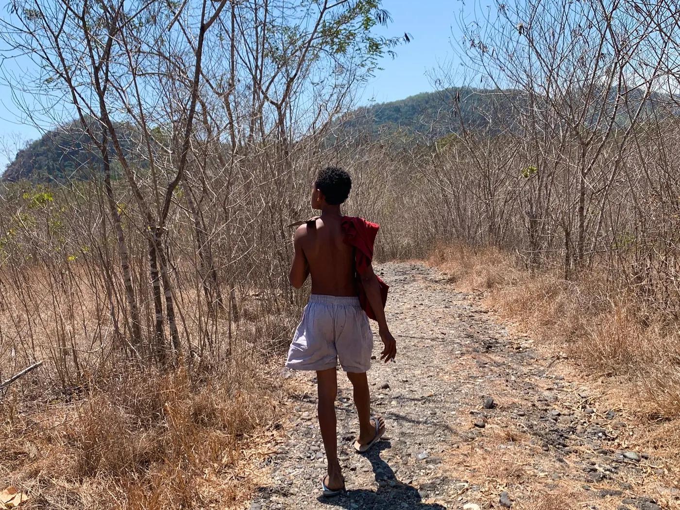 A young man walking down a dry forest trail under the bright sun, showcasing a natural landscape.