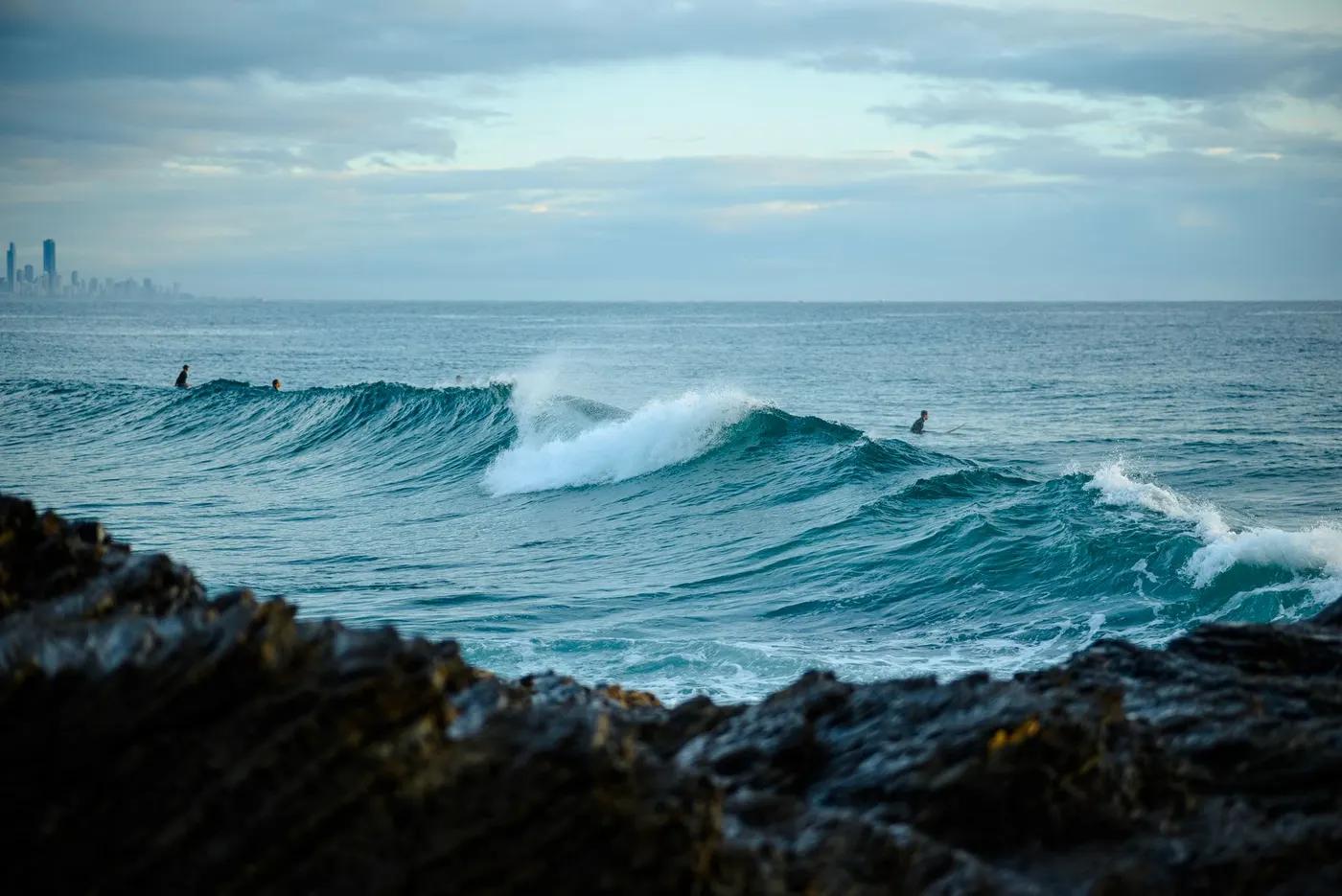 Surfers catching waves on Currumbin Beach with rocky shoreline and distant city skyline.
