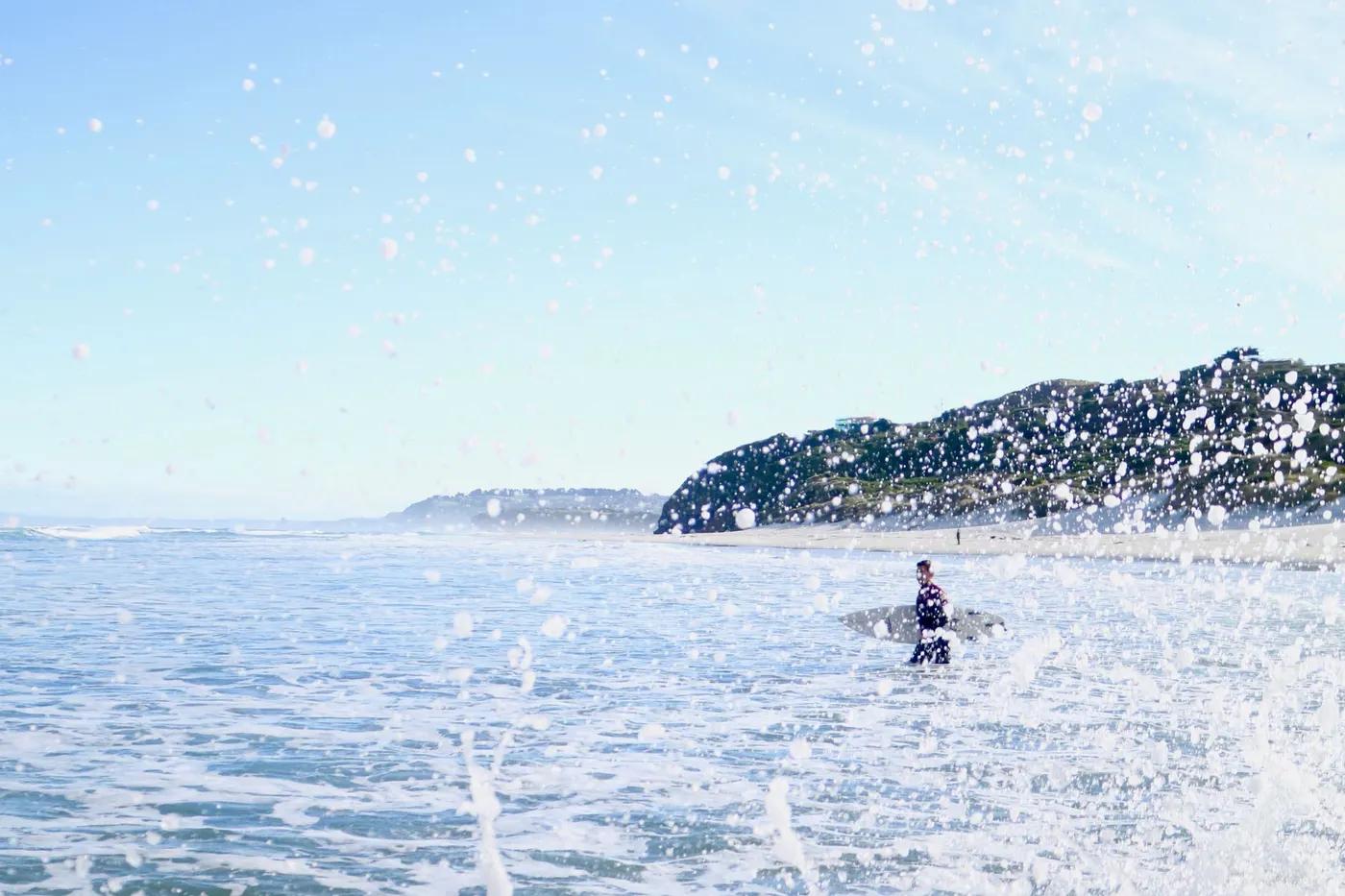 A surfer catching waves amidst ocean spray on Dunedin Beach, New Zealand.