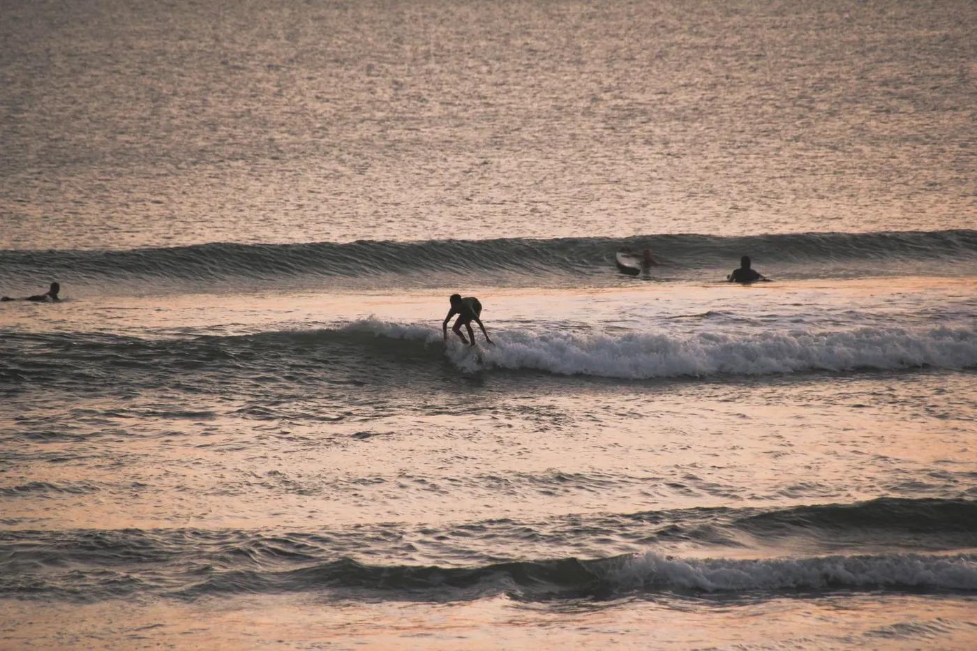 A surfer rides the waves during a stunning sunset on a Bali beach, showcasing water sports excitement.