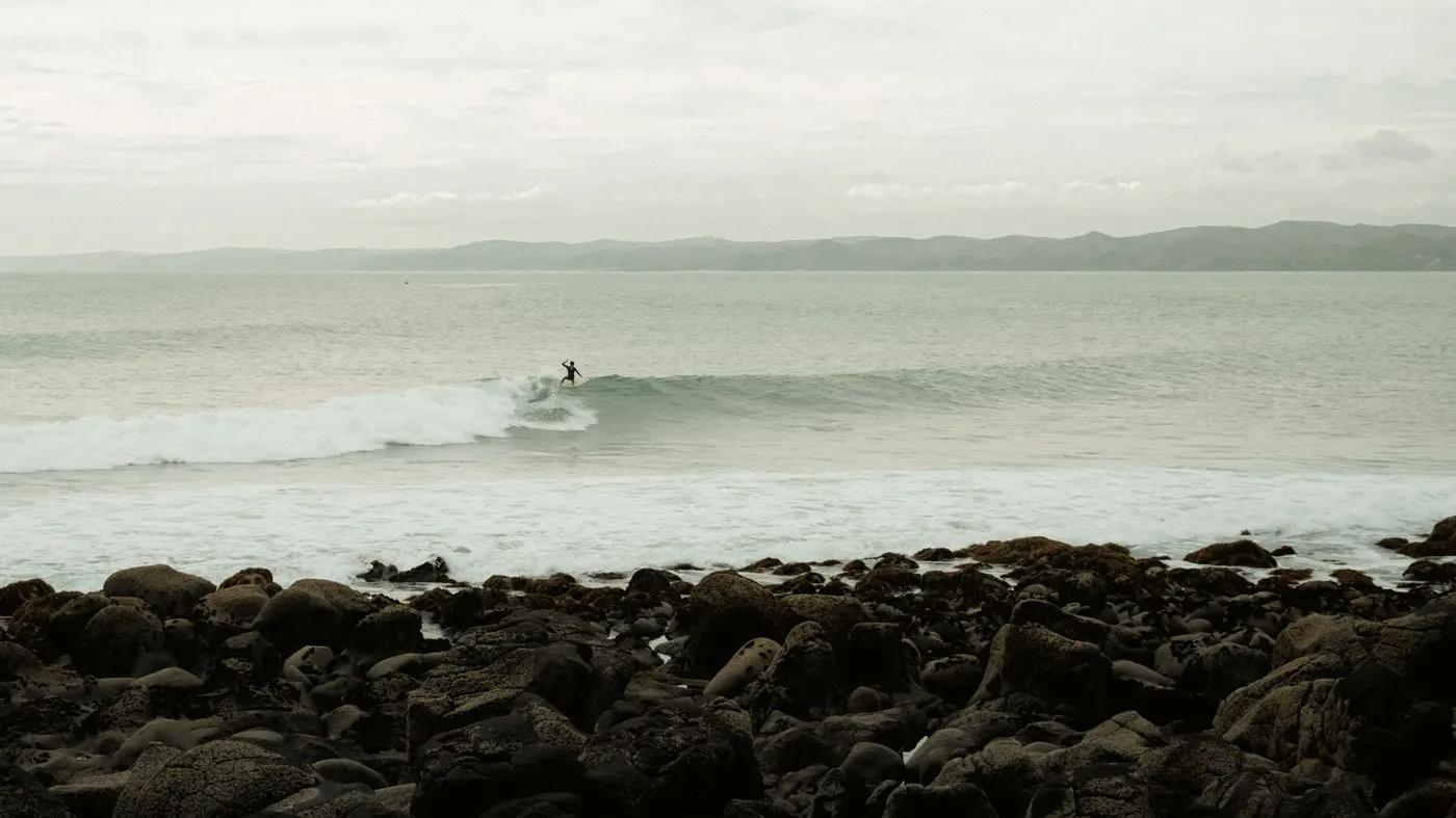 A surfer catches a wave on the scenic Raglan Beach in New Zealand, with rocky shorelines and distant hills.