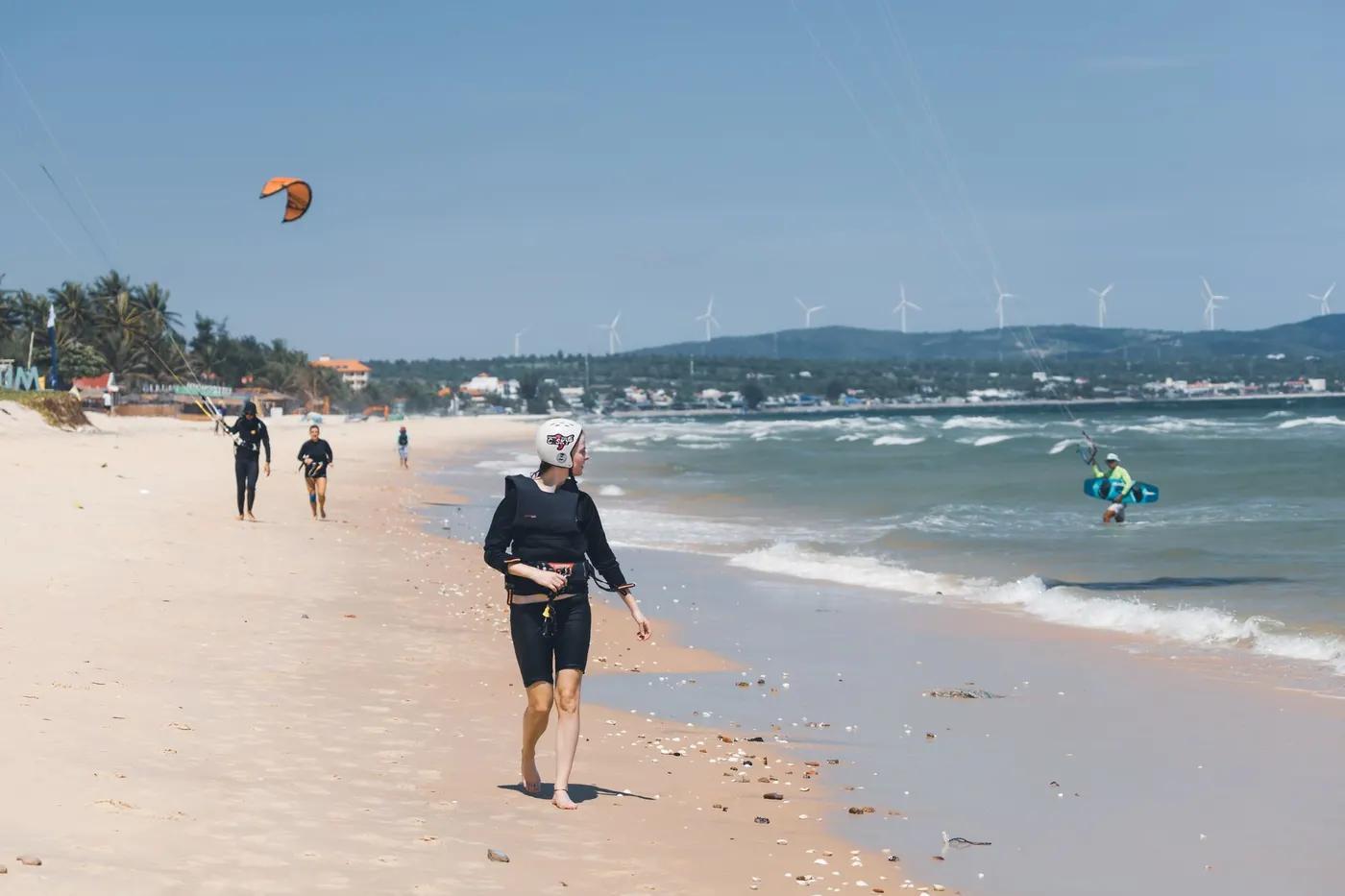 Kitesurfers walk and surf on a sunny beach with wind turbines in the background, capturing a vibrant seaside activity s…